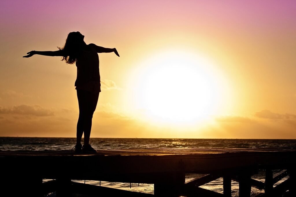 Silhouette of woman standing on a dock beside a large body of water with outstretched arms in front of a sunset